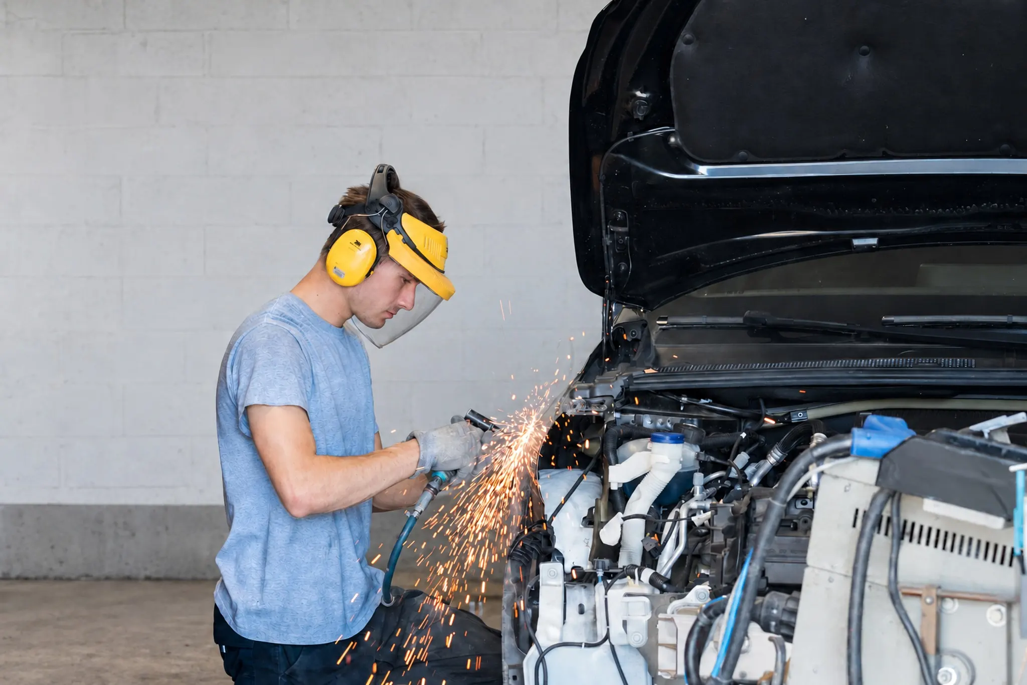 Firmenfotografie: Mitarbeiter mit Gehörschutz und Gesichtsschild schleift in der Werkstatt an einem Auto, Funken sprühen