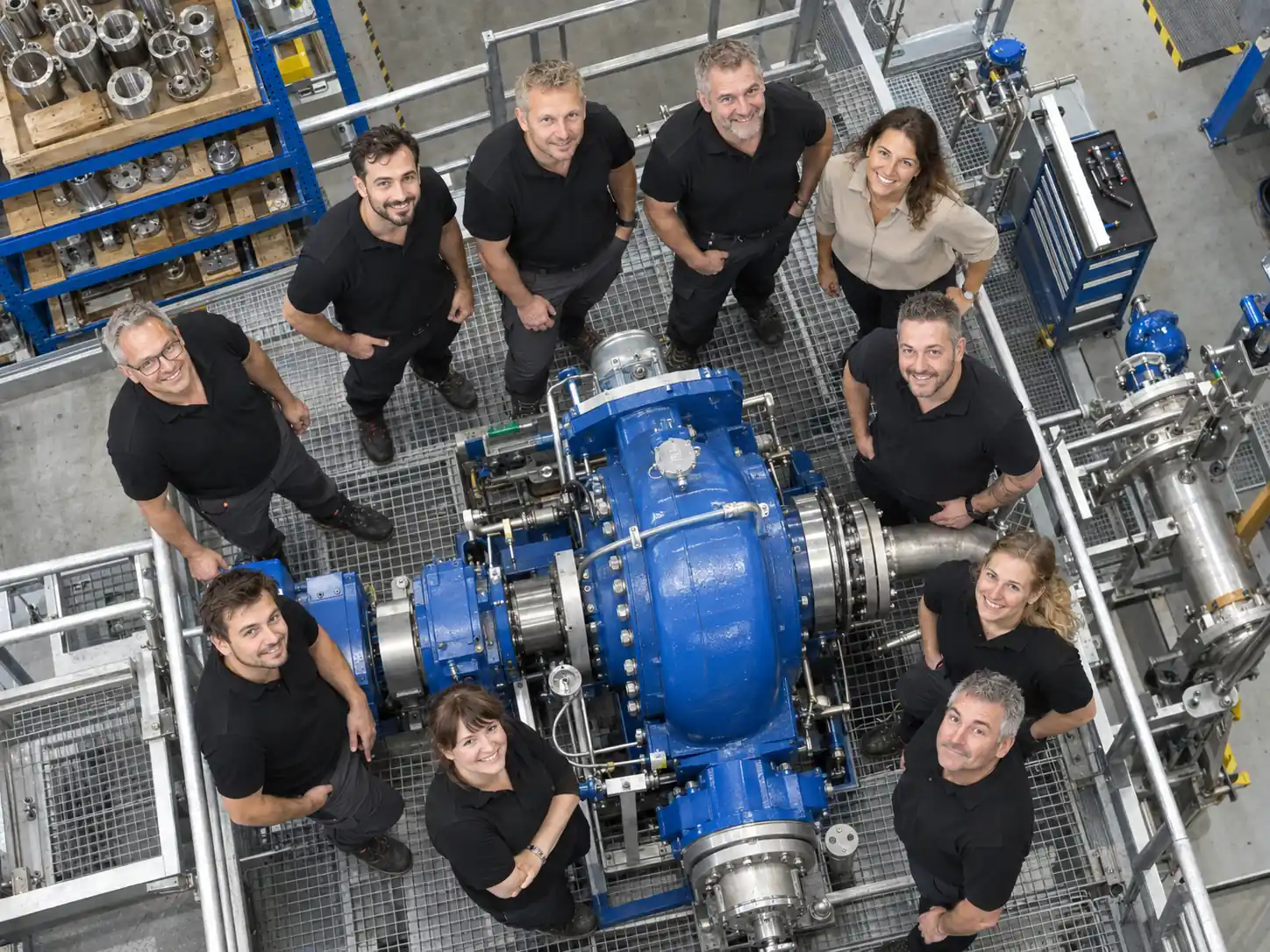 Teamfoto von oben: zehn Mitarbeitende stehen in der Werkhalle um eine blaue Pumpanlage